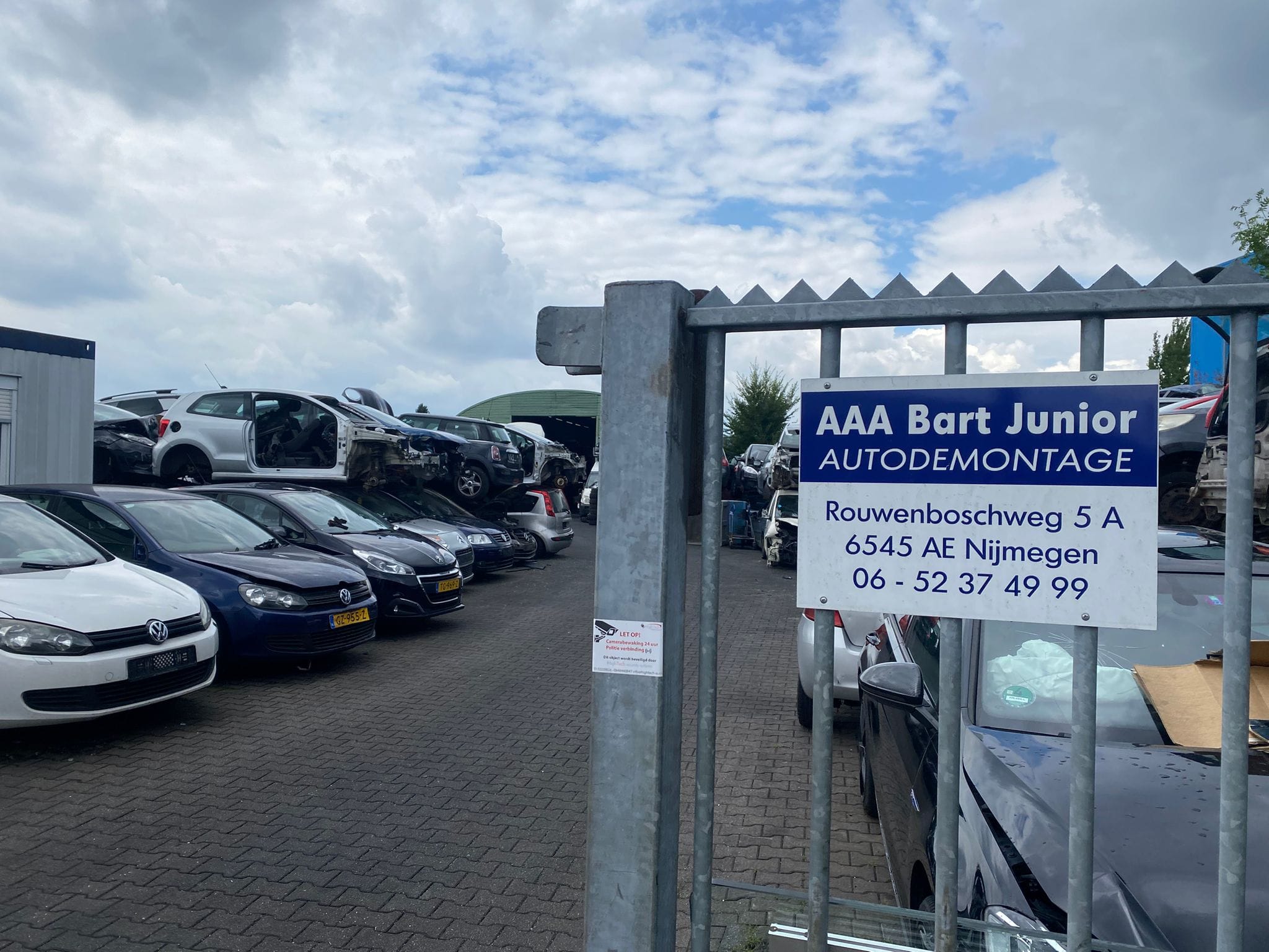 Ingang van Autosloperij Bart Junior in Nijmegen. Op de voorgrond is een metalen hek te zien met een wit bedrijfsbord, en daarachter een terrein met rijen geparkeerde en gestapelde sloopauto's