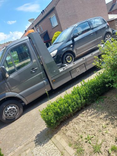Grijze autotransporter met een zwarte sloopauto op de laadklep, geparkeerd in een woonwijk in Rijkerswoerd. Gratis ophaalservice voor sloopauto’s met directe uitbetaling